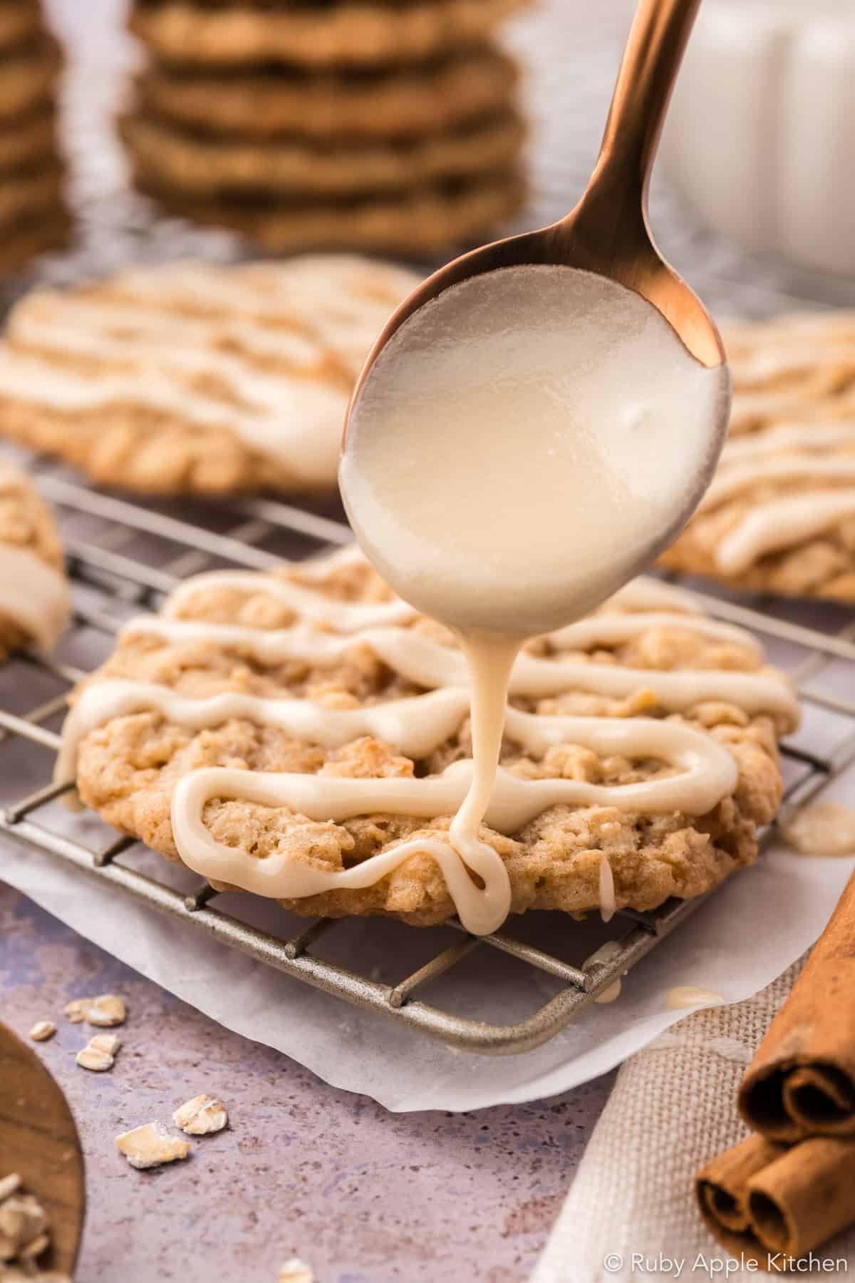Maple glaze pouring from a spoon onto an oatmeal cookie, with more glazed cookies nearby.