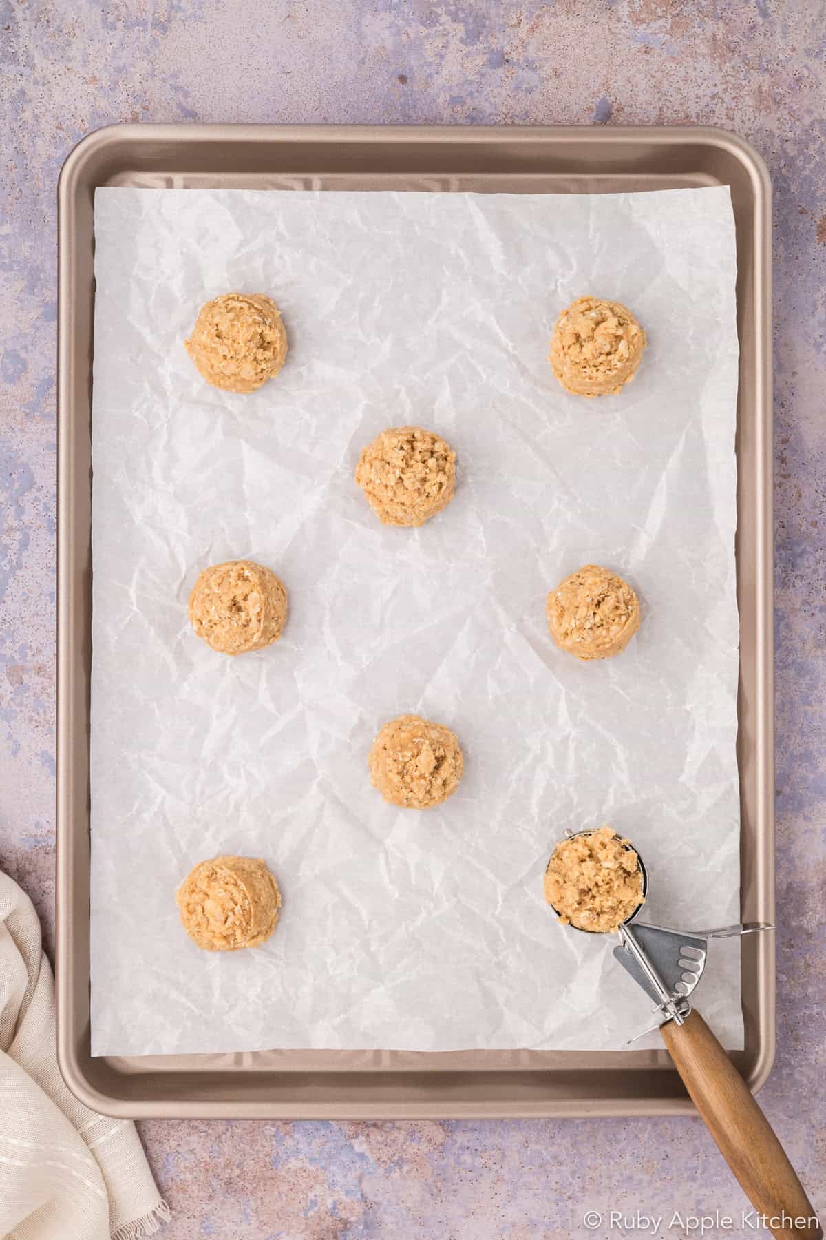 Rounded portions of maple oatmeal cookie dough spaced on a parchment-lined baking sheet, with a cookie scoop in the corner.